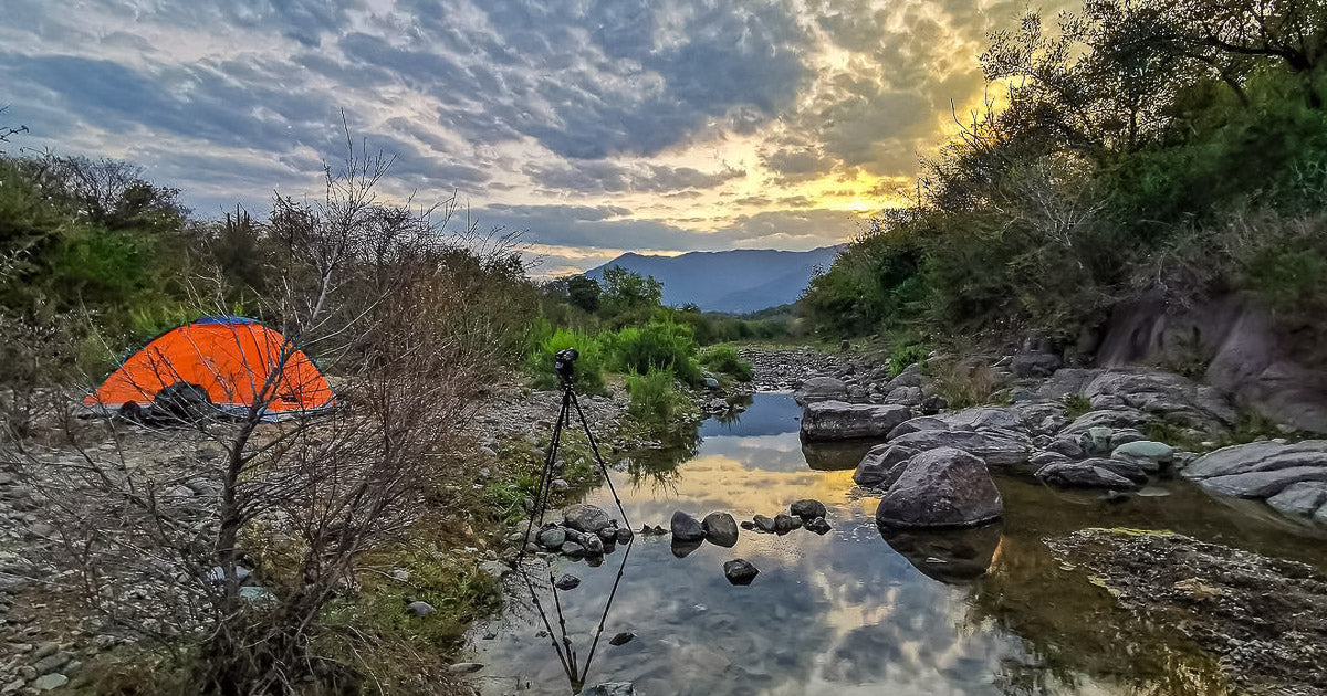 Tienda de campaña naranja junto a un río pedregoso al amanecer, con un trípode y montañas reflejadas en una poza.