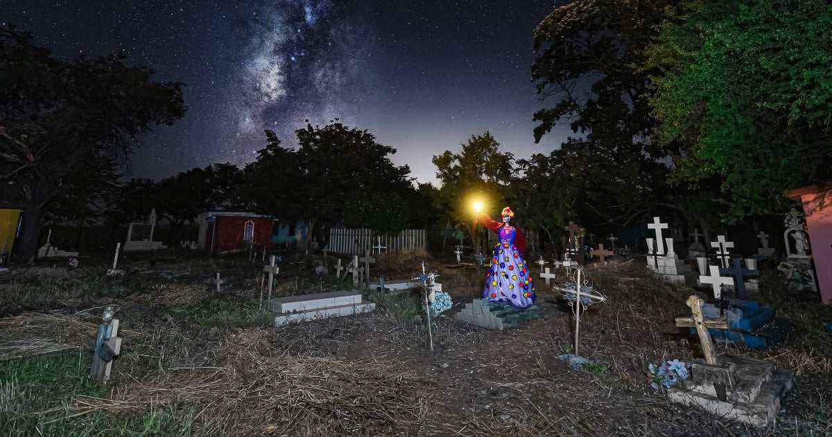 Catrina con vestido colorido iluminando con una linterna en un panteón rural, rodeada de cruces y tumbas bajo un cielo nocturno dominado por la Vía Láctea.