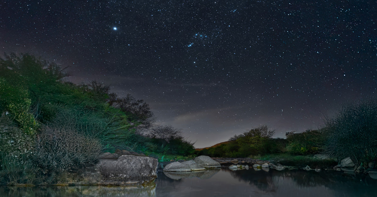 La constelación de Orión destaca en un cielo estrellado sobre un río tranquilo con rocas y matorrales.