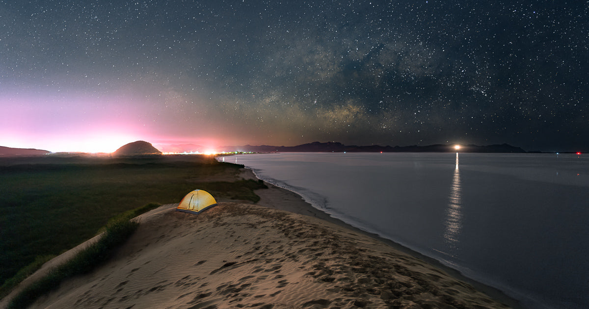 Tienda de campaña iluminada sobre una duna a la orilla del mar; cielo estrellado con la Vía Láctea y luces urbanas en el horizonte.