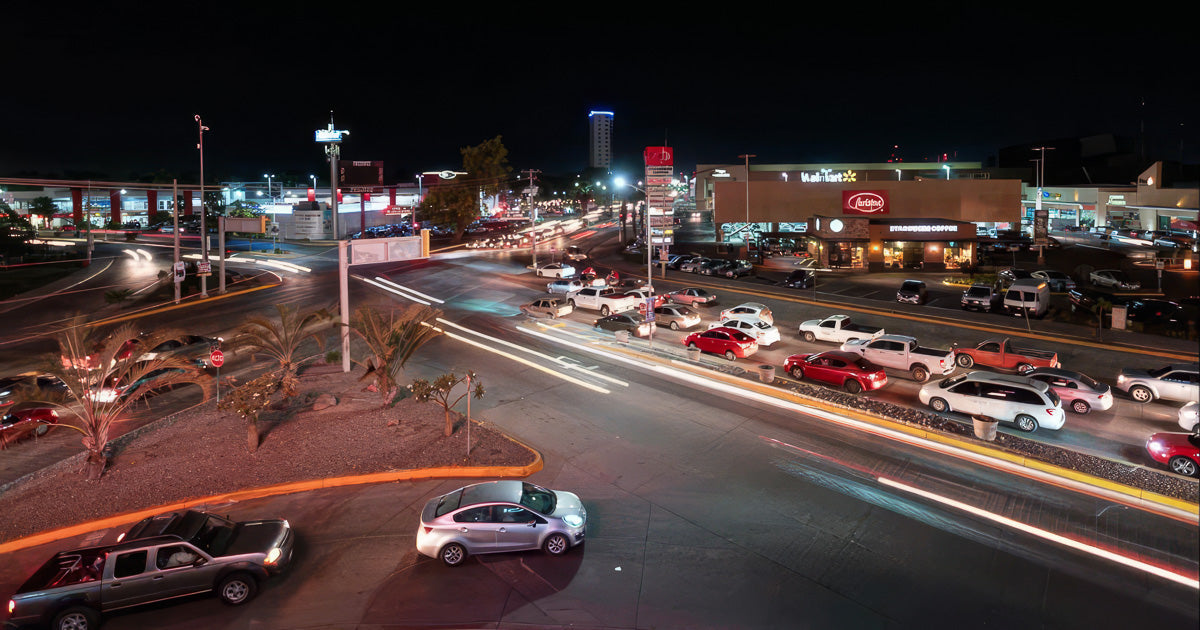 Cruce vial de noche con estelas de luz por larga exposición, fila de autos junto a una plaza comercial y cielo oscuro.