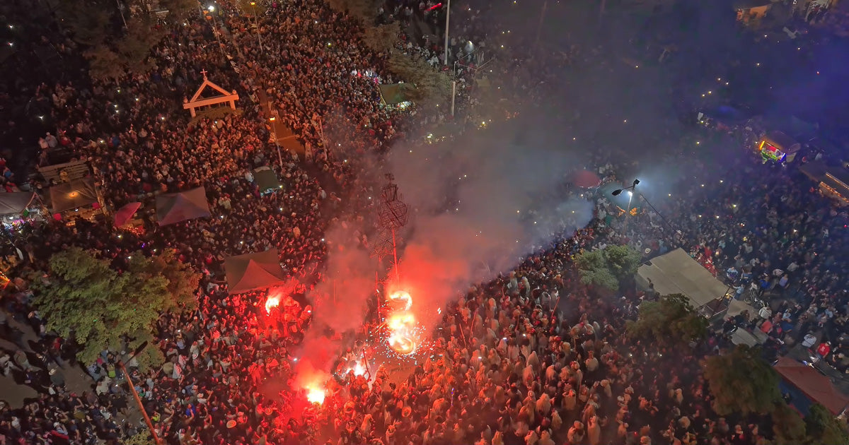 Multitud vista desde dron durante el encendido del torito de fuegos artificiales en la plaza; humo y bengalas rojas rodean la estructura.