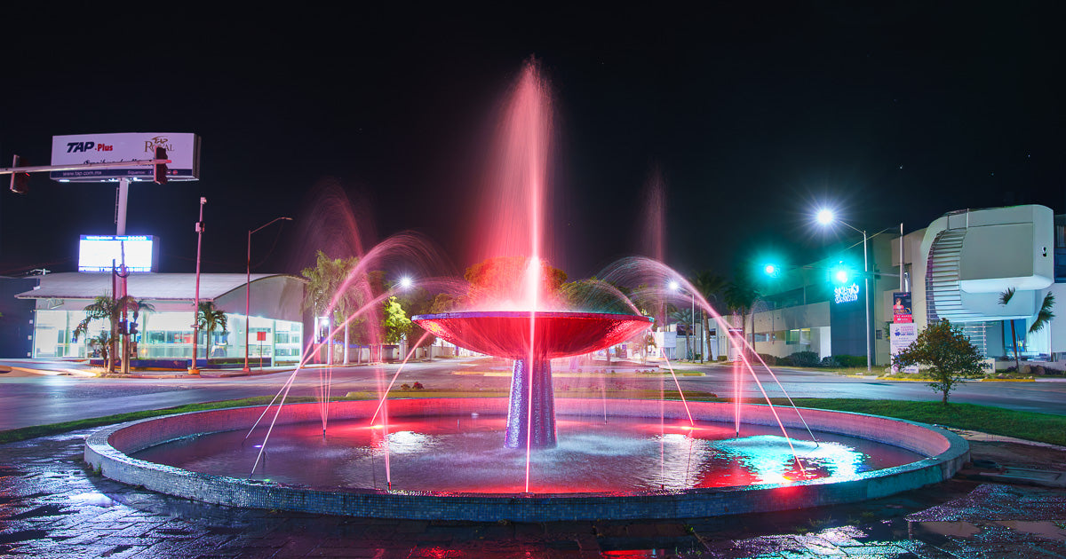 Fuente circular iluminada en rojo con chorros y luces nocturnas en una glorieta urbana; tráfico y edificios al fondo.