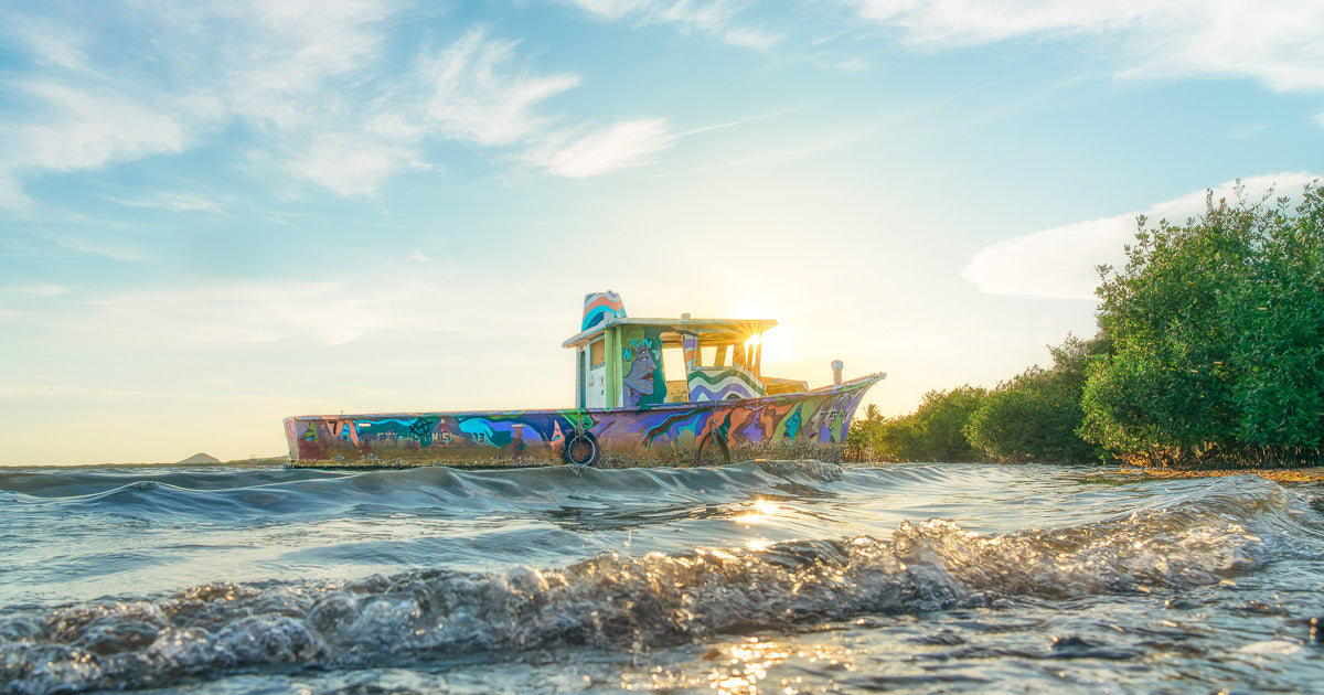 Embarcación pequeña con mural multicolor, varada en agua somera junto a mangle; sol a contraluz al atardecer, olas suaves en primer plano y cielo claro.