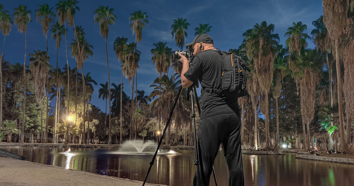 Fotógrafo con trípode junto a un estanque con fuente, rodeado de palmeras altas y luminarias encendidas; cielo nocturno, toma a ras de suelo.