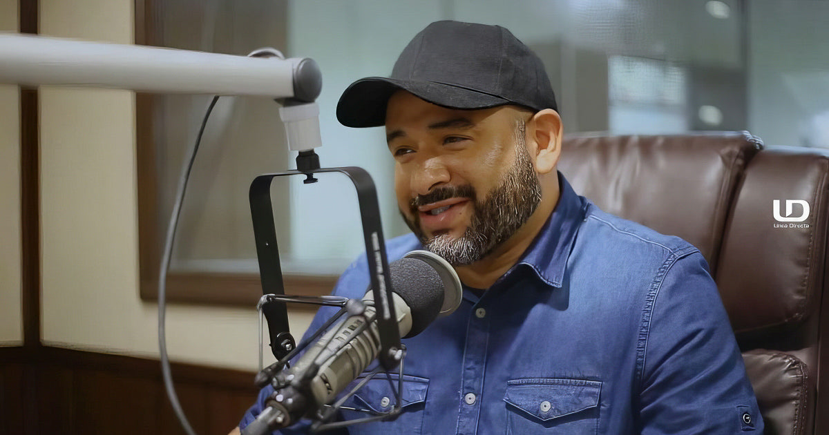Hombre con gorra negra y camisa de mezclilla habla sonriente frente a un micrófono de estudio, sentado en cabina de radio con ventanal y luz suave.