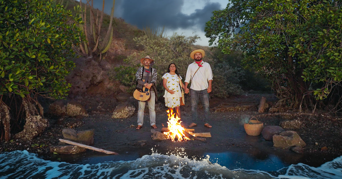 Tres personas de pie junto a una fogata encendida en la orilla, rodeadas por manglar y cerro al atardecer, con el oleaje entrando al cuadro y una canasta sobre la arena húmeda.