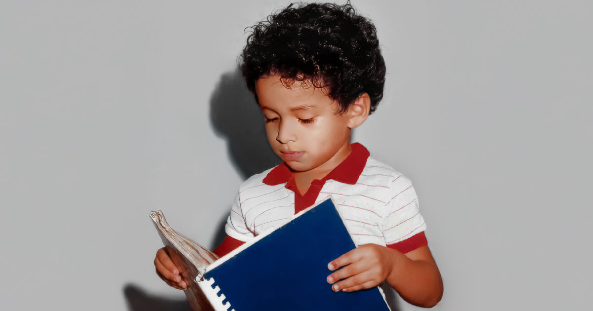 Niño de cabello rizado sostiene y hojea un cuaderno azul de pasta dura, concentrado, en interior con fondo gris liso y luz frontal suave.