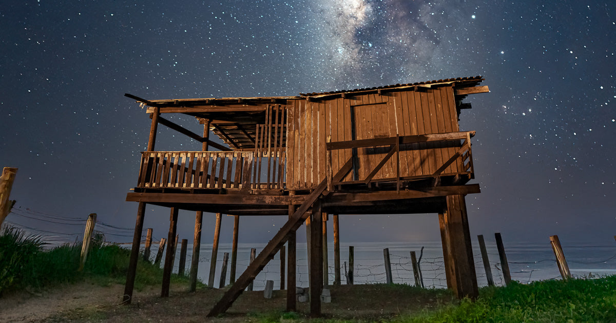 Palafito de madera frente al mar, con escaleras y barandal, rodeado de postes y alambre; cielo nocturno con Vía Láctea visible, toma a ras de suelo.