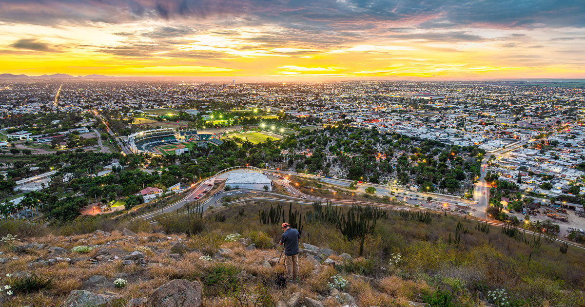 Panorámica de Los Mochis al atardecer desde el Cerro de la Memoria, con el estadio de los Cañeros iluminado y la ciudad extendiéndose hasta el horizonte.