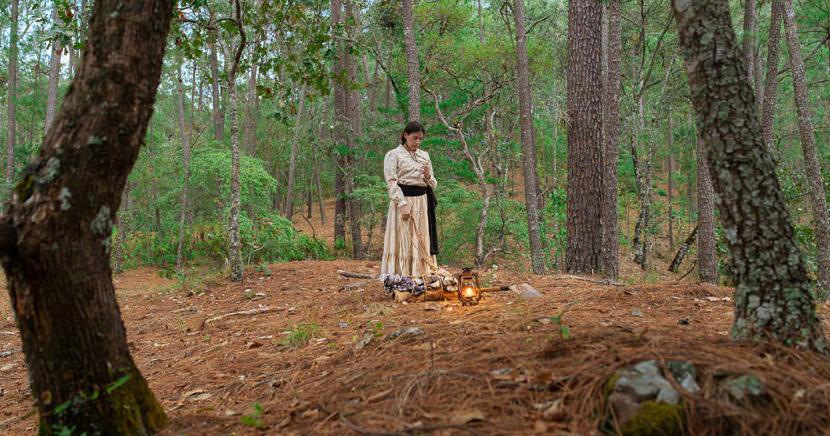 Mujer con vestido antiguo de tono claro en un bosque de pinos, junto a un farol de aceite encendido y un fardo en el suelo cubierto de hojas y agujas.
