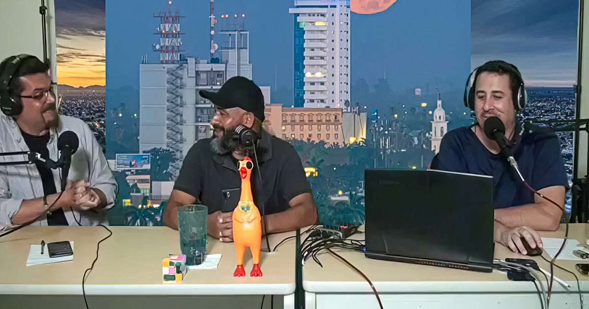 Tres conductores conversan en un estudio de podcast con micrófonos y laptop; al centro hay una gallina de hule y, de fondo, un gran cartel de la luna llena sobre Los Mochis.