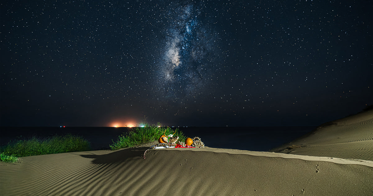 Noche estrellada con la Vía Láctea sobre una duna; en primer plano una ofrenda con máscara de venado, sonajas de calabaza y manta sobre la arena junto al mar.