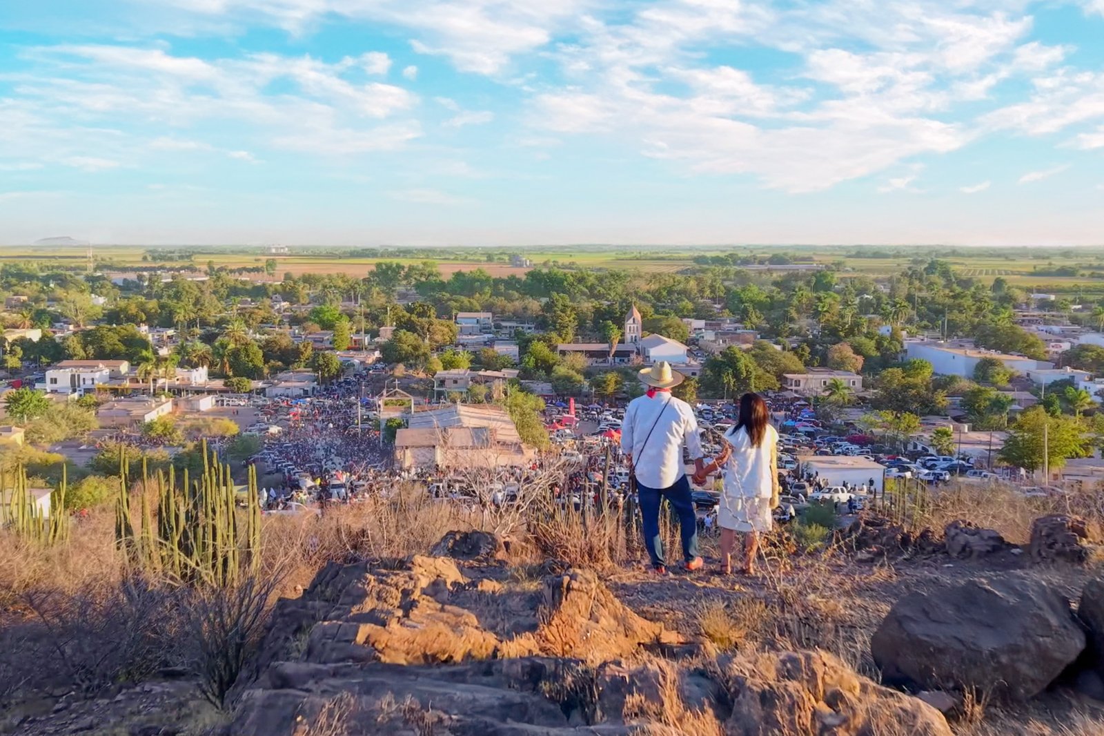 Vista panorámica del pueblo desde el cerro; dos personas de espaldas observan la plaza llena de gente.