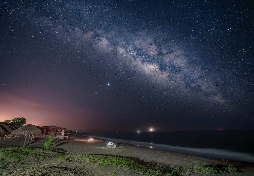 Arco de la Vía Láctea sobre playa con palapas y dos tiendas de campaña iluminadas; mar en calma y luces lejanas en el horizonte nocturno.