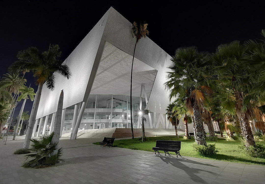 Edificio de fachada blanca con volúmenes angulares, palmeras y bancas; escena nocturna con iluminación lateral.