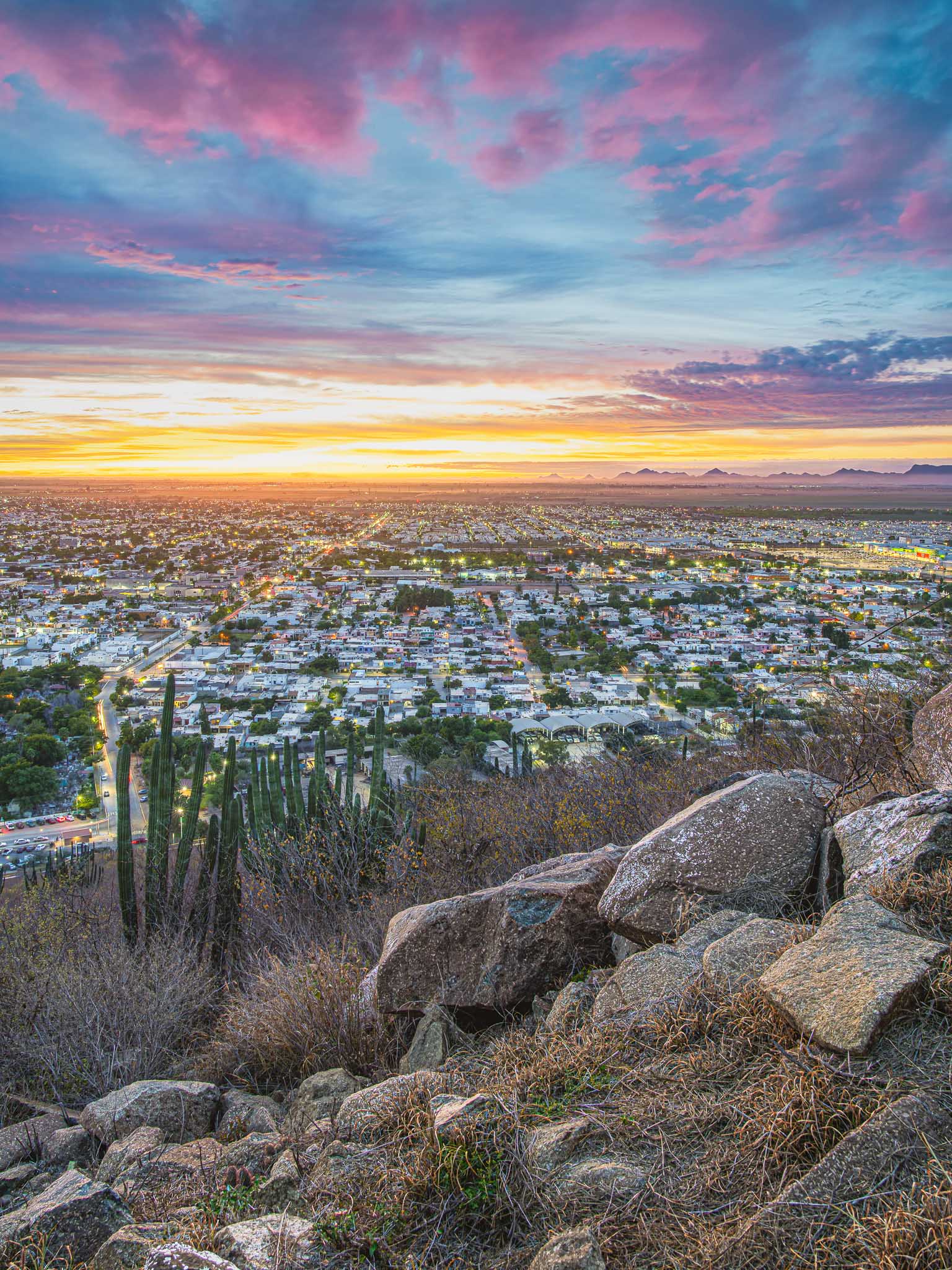 Vista vertical desde ladera rocosa con cactus; ciudad iluminada al crepúsculo, nubes rosadas y montañas lejanas.