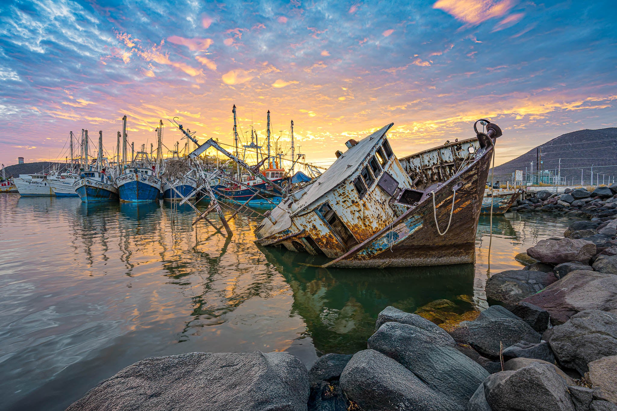 Barco camaronero oxidado inclinado y semihundido en muelle; lanchas al fondo, rocas en primer plano, amanecer colorido.