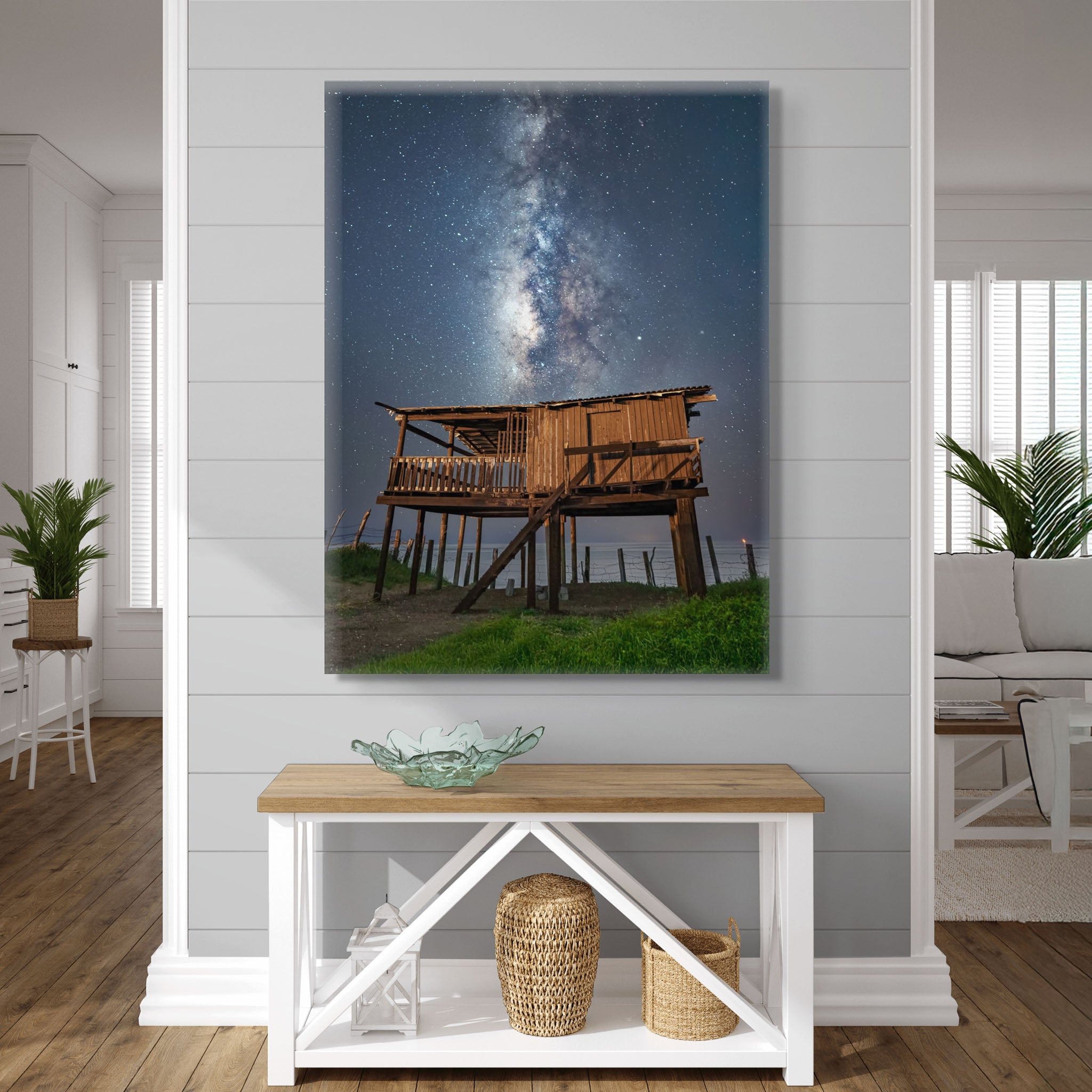Canvas colgado en interior claro; casa de madera elevada en primer plano bajo cielo estrellado; atmósfera nocturna, tonos neutros y sensación de calma.