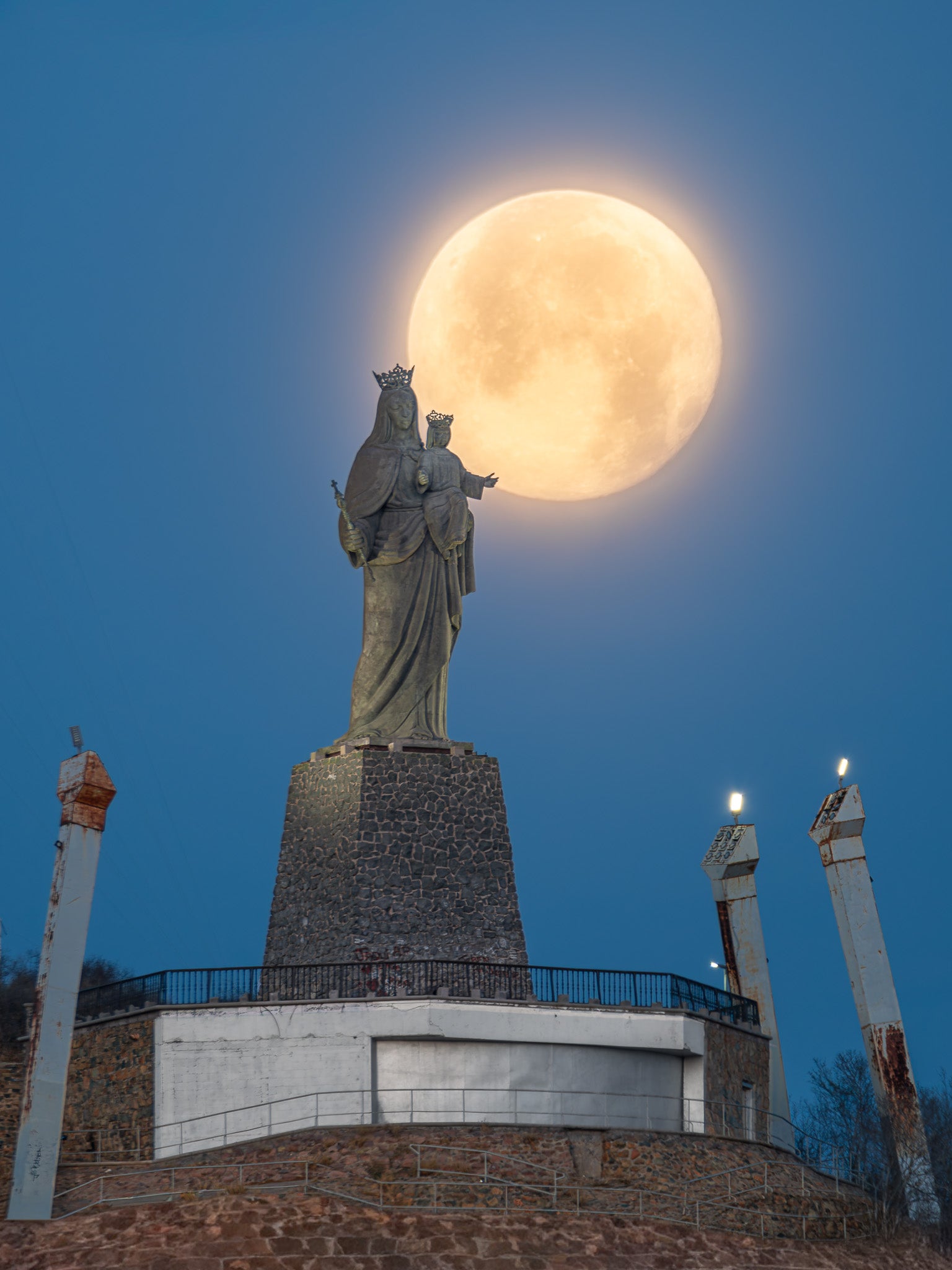 Escultura de la Virgen con el Niño sobre pedestal de piedra en hora azul; luna llena detrás forma un halo suave en el cielo.