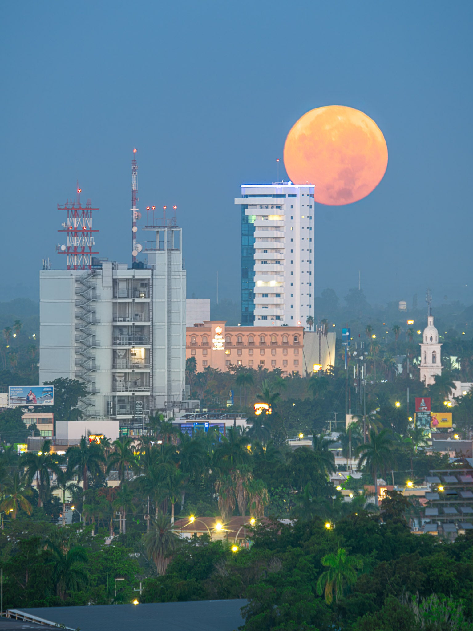 Luna llena anaranjada sobre horizonte urbano; torres y antenas entre palmeras, luces tenues de amanecer, vertical.