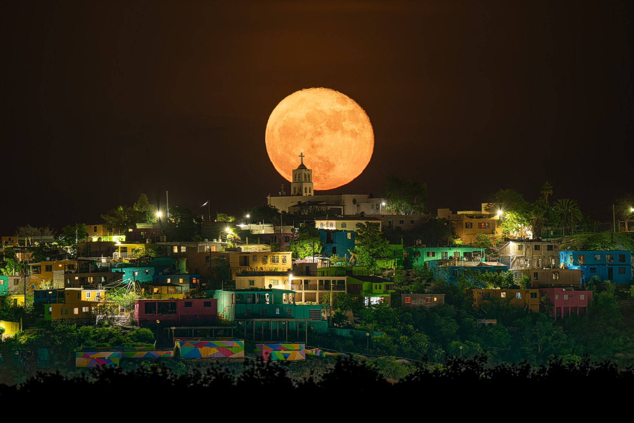 Superluna naranja alineada sobre torre de iglesia; barrio en ladera con casas coloridas iluminadas y siluetas de árboles en la noche.