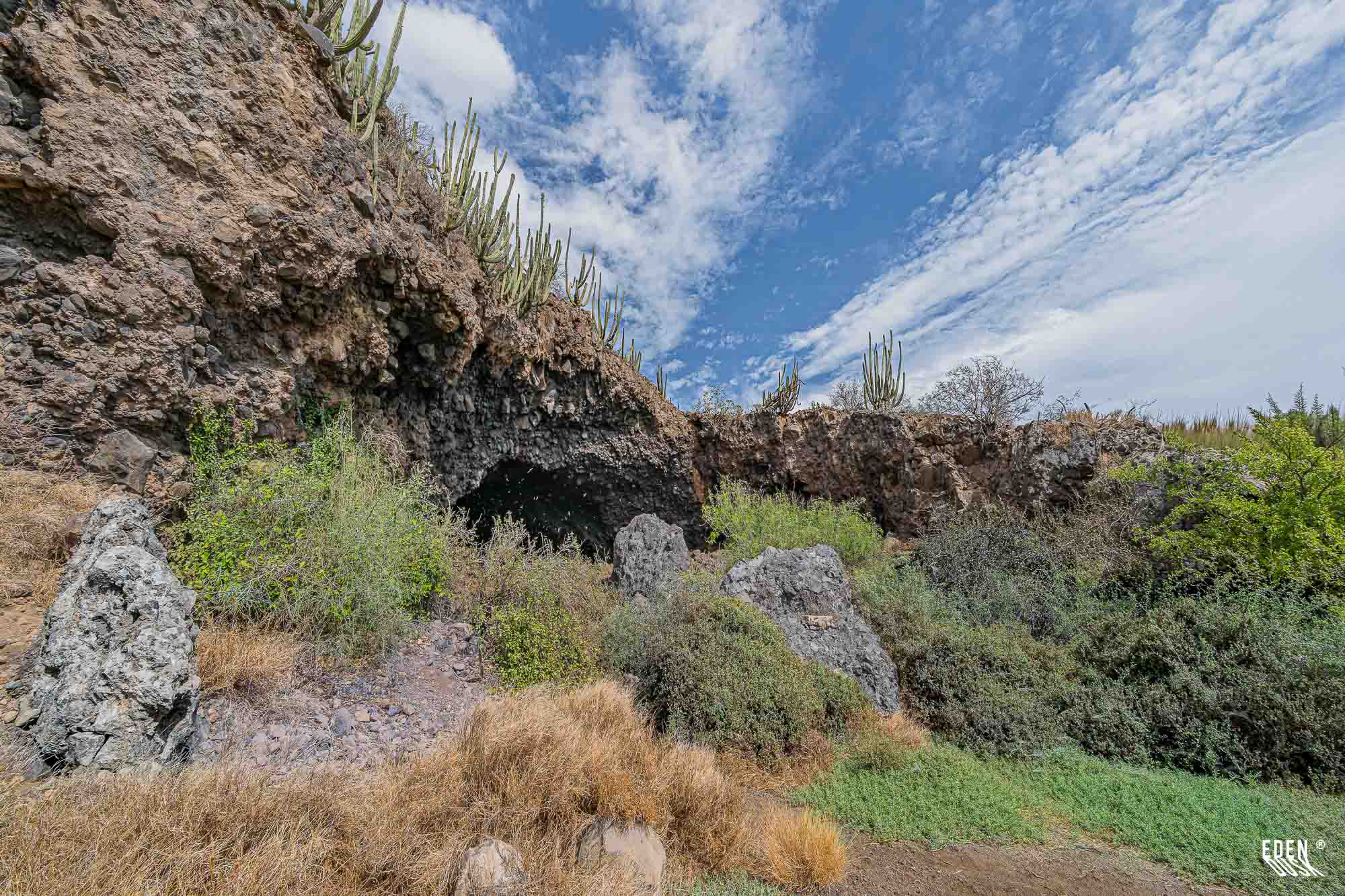 Entrada rocosa de la Cueva de los Murciélagos con cactus sobre la ladera y cielo azul con nubes, El Maviri, Sinaloa.