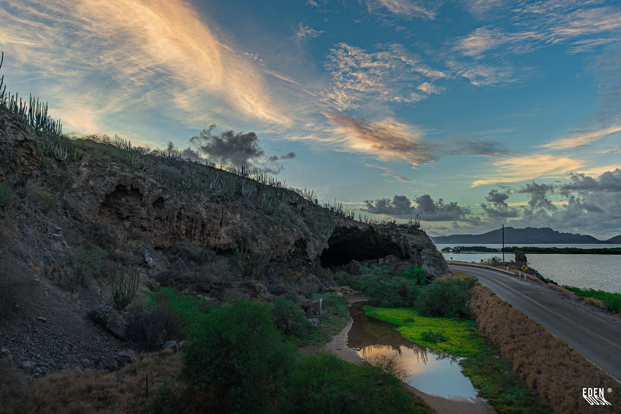 Entrada de la Cueva de los Murciélagos al atardecer con nubes encendidas, carretera contigua y charcos entre matorrales junto a la laguna, El Maviri, Sinaloa.