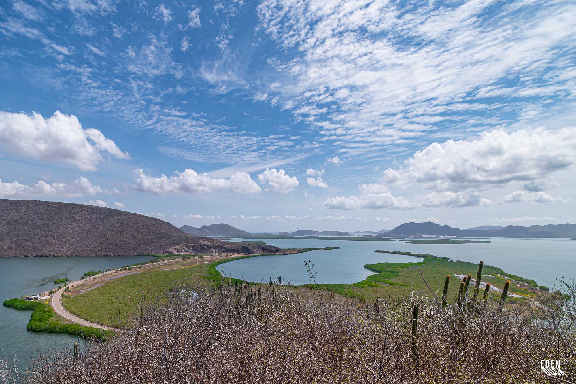 Vista panorámica desde la Cueva de los Murciélagos con lagunas y manglares, carretera curva y sierras al fondo bajo cielo con nubes, El Maviri, Sinaloa.