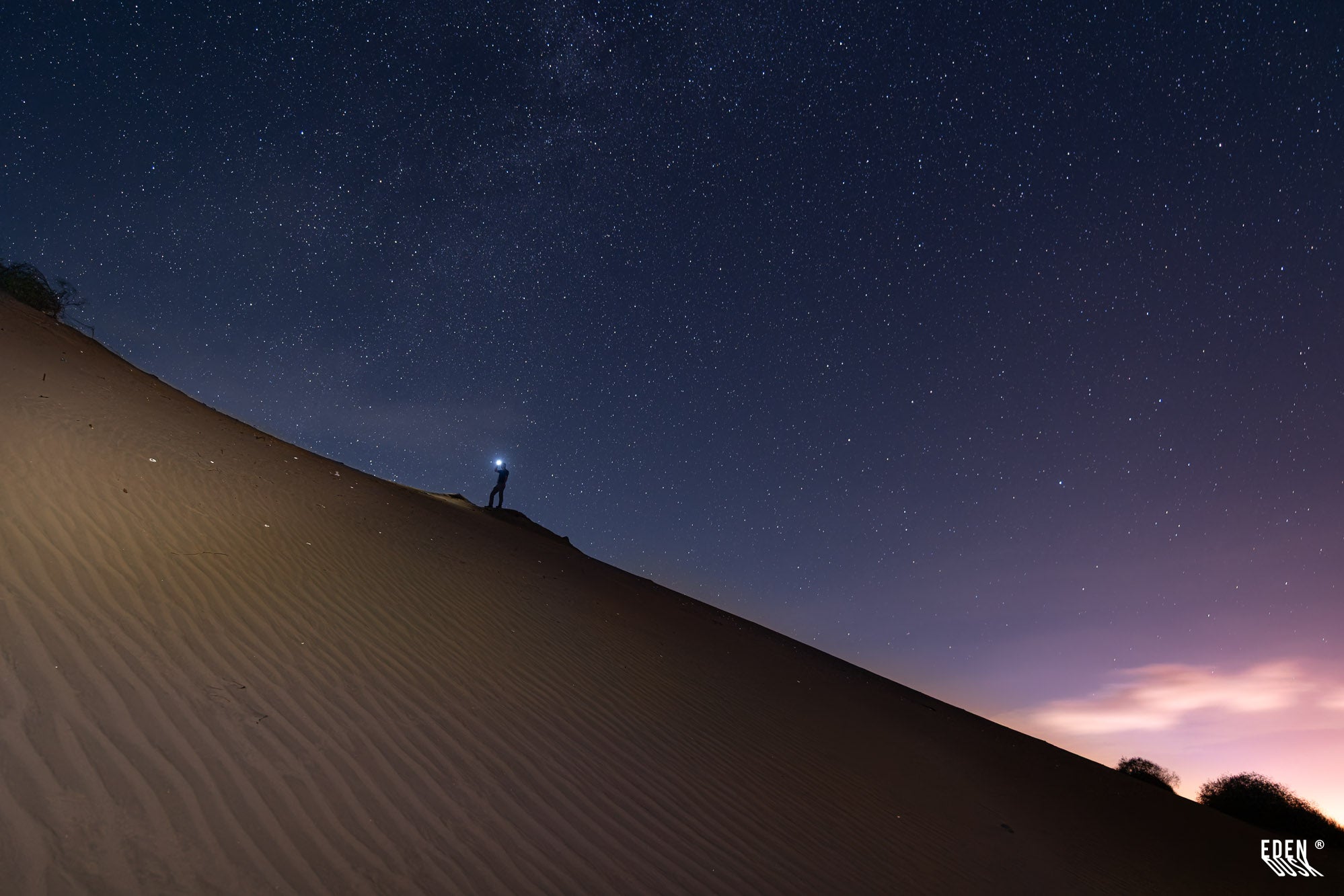 Ladera de duna con estrías en primer plano; silueta de persona con luz en la cresta bajo cielo nocturno estrellado y nubosidad tenue.