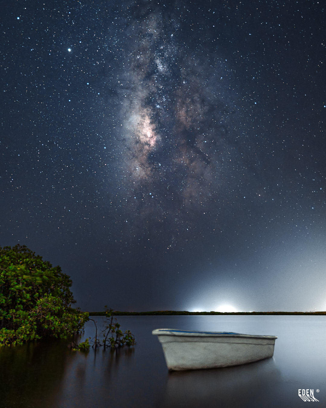 Lancha blanca flotando en una laguna tranquila junto a manglar, con la Vía Láctea brillante y un resplandor distante en el horizonte, El Maviri, Sinaloa.