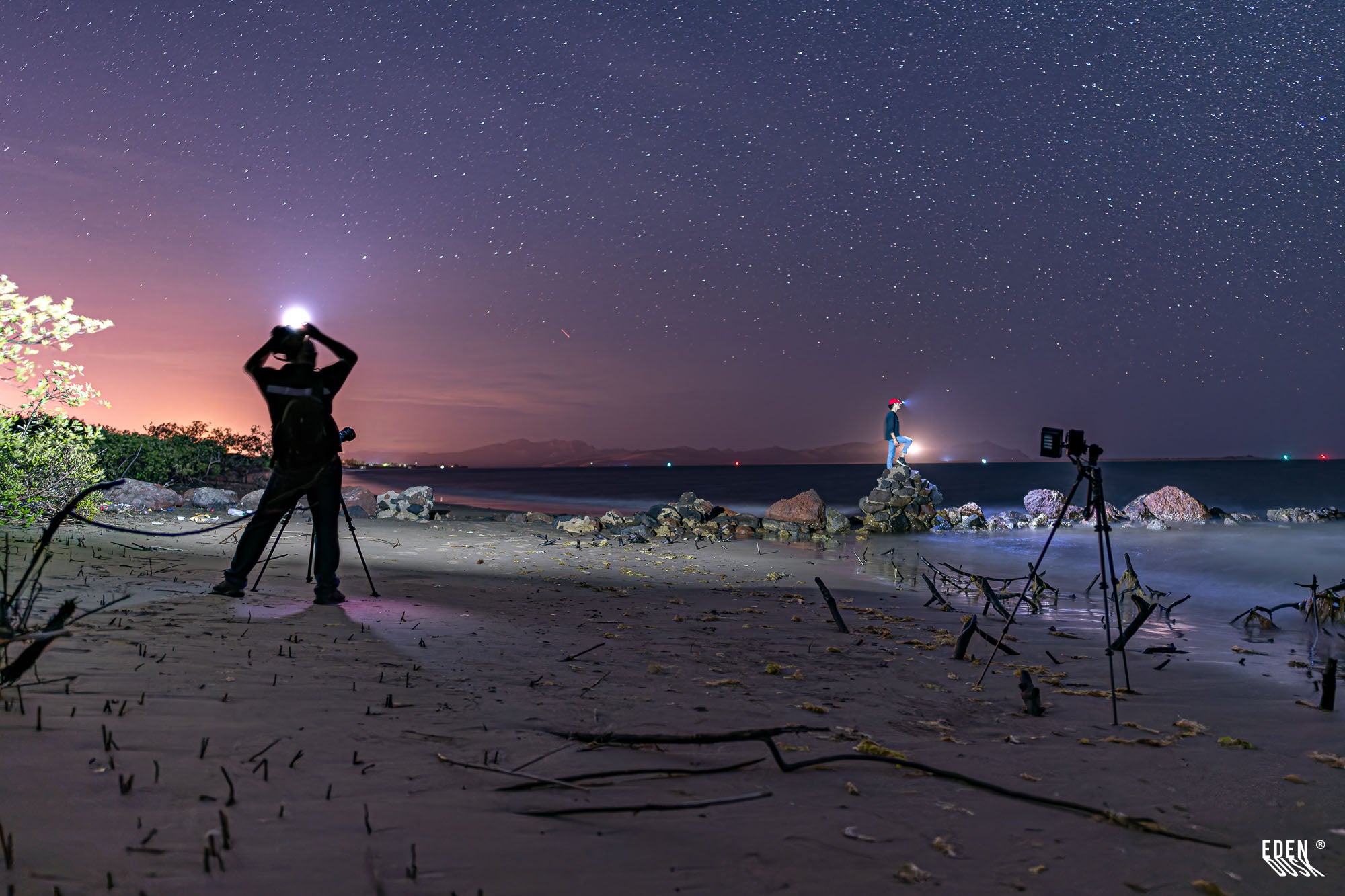 Playa nocturna con dos personas iluminadas con linternas en la cabeza, trípodes y rocas; cielo estrellado y mar en calma.