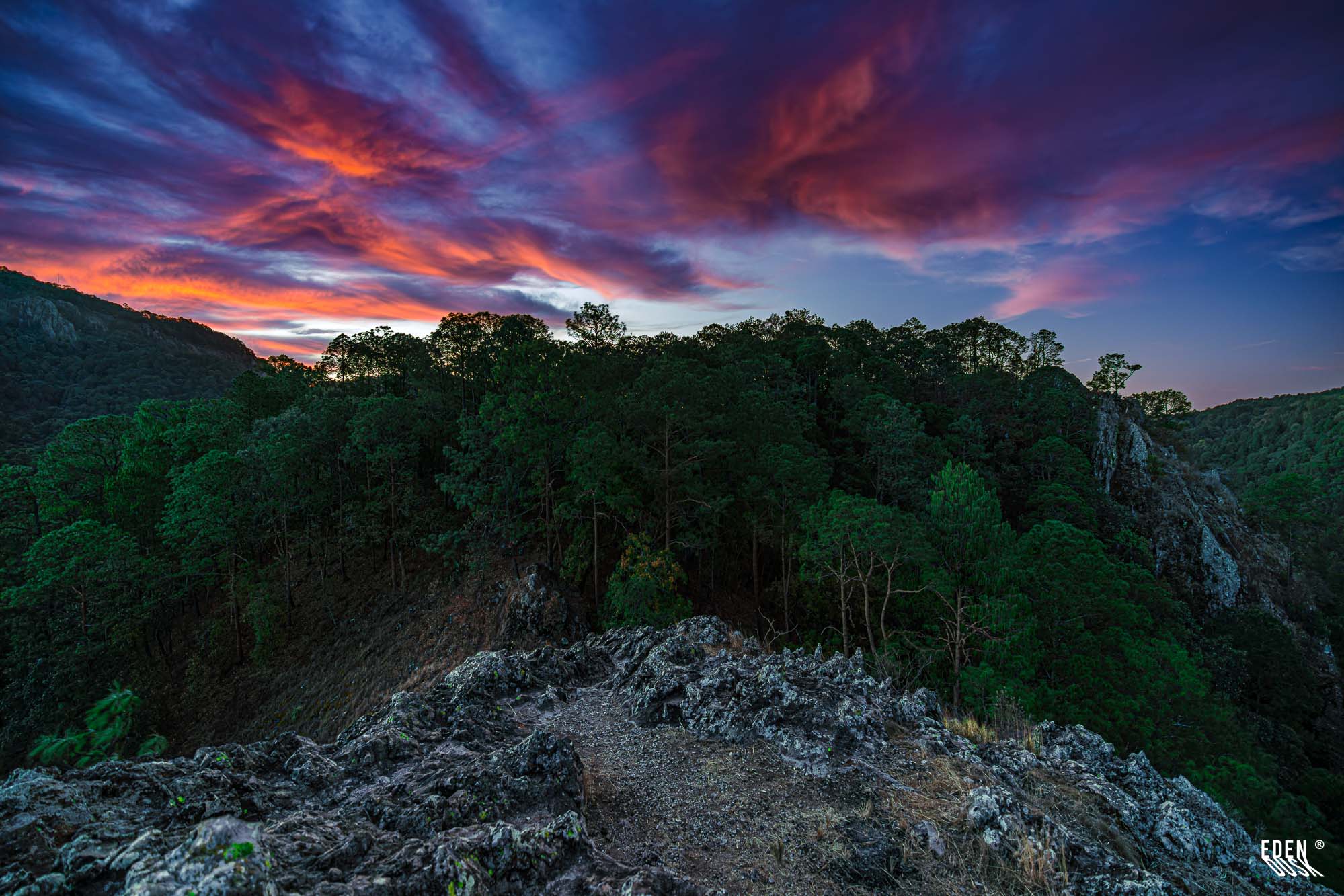 Cresta rocosa en primer plano frente a un bosque denso; cielo del atardecer con nubes rojizas y azul profundo.