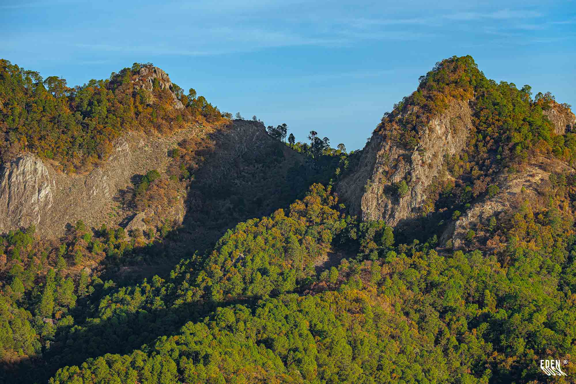 Dos cerros rocosos con laderas cubiertas de árboles; sombra marcada en el valle central y cielo azul despejado.