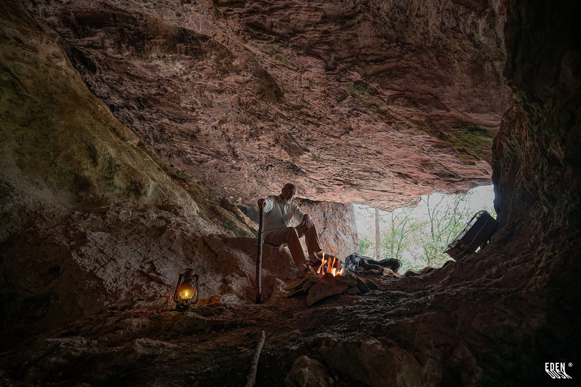 Hombre sentado junto a una fogata dentro de una cueva; lámpara encendida a la izquierda y entrada rocosa con árboles al fondo.