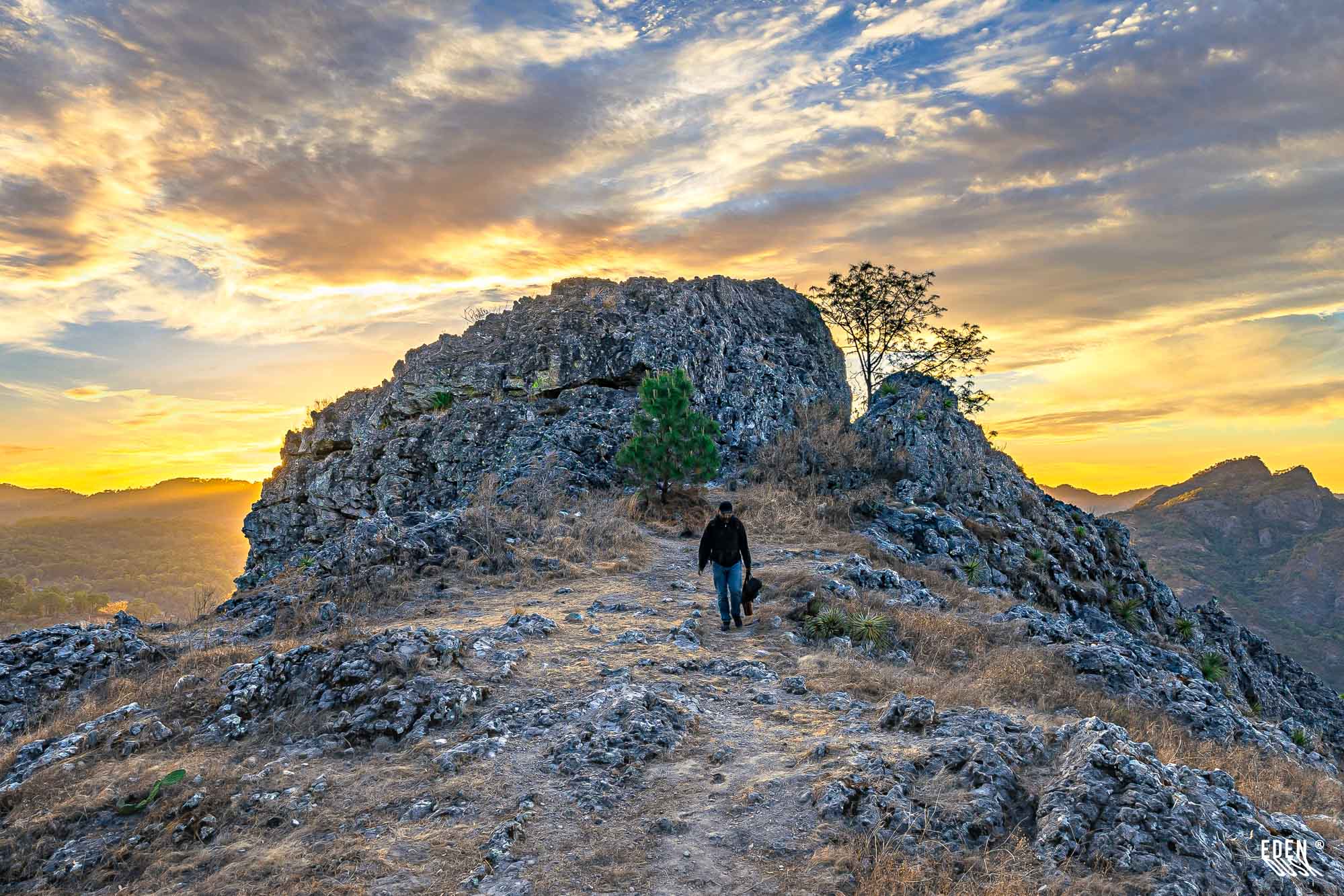 Persona en sendero rocoso frente a loma de piedra; cielo de atardecer con nubes doradas y valle a los lados.