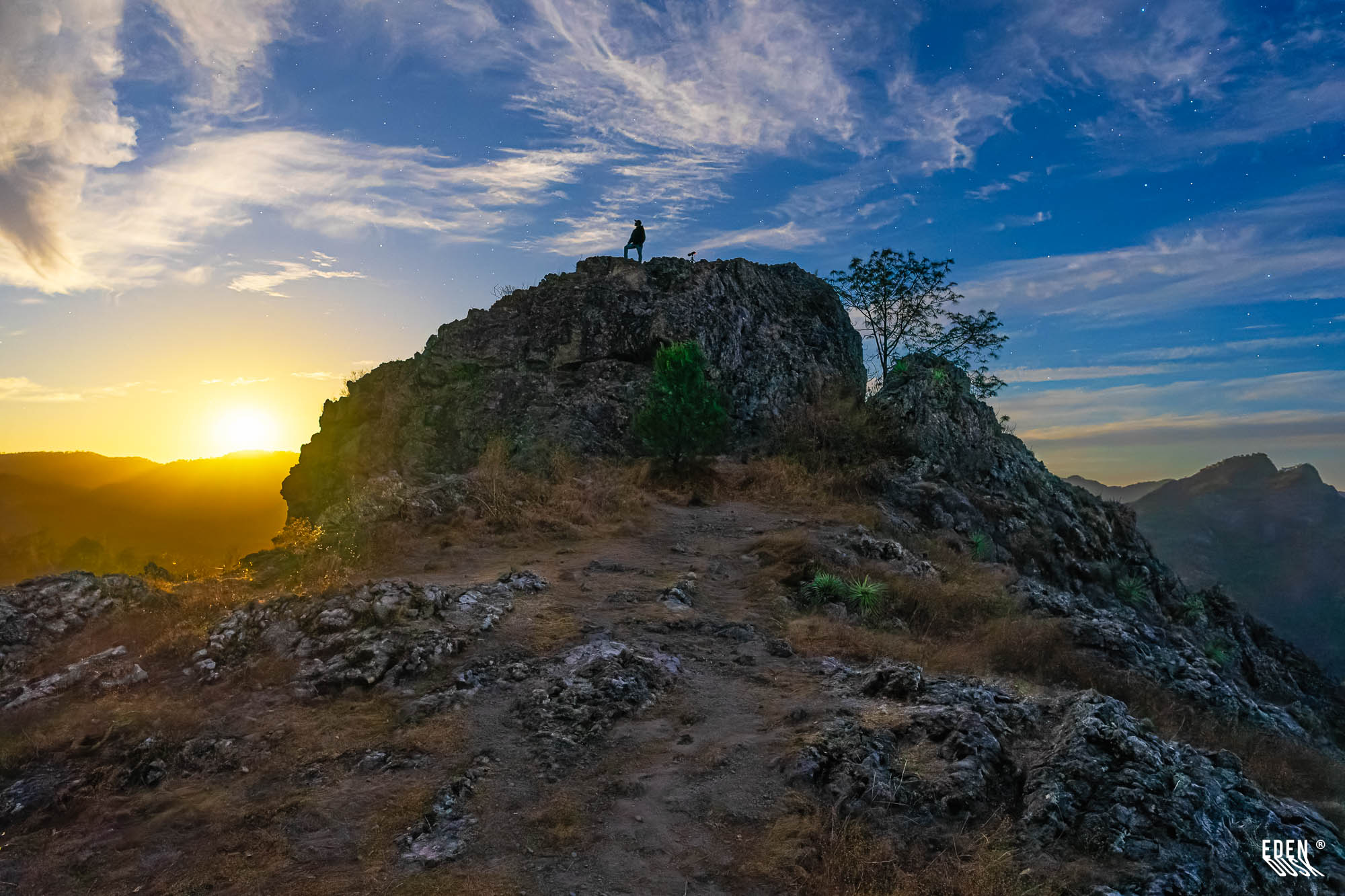 Persona de pie en la cima de un peñasco; sol bajo a la izquierda, cielo azul con nubes y algunas estrellas visibles.