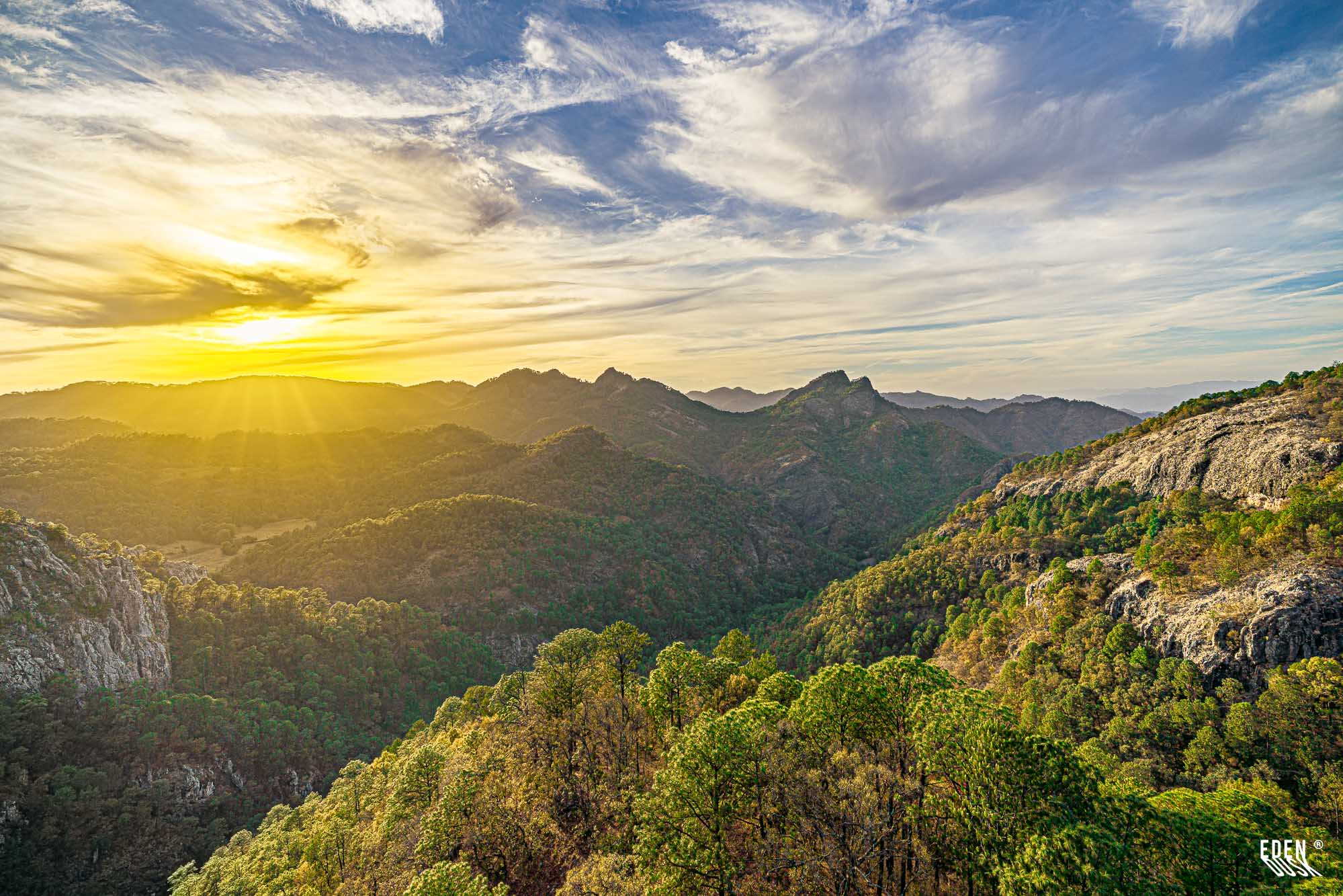 Sol bajo tras la sierra con rayos abiertos sobre valles arbolados; laderas rocosas a la derecha y cielo con nubes altas.